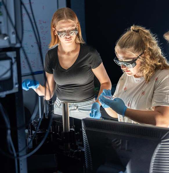 Students wearing safety glasses and gloves conducting laboratory experiment with equipment and glassware under focused lighting.