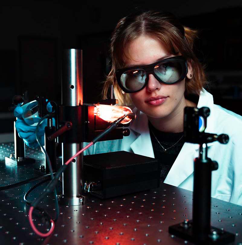 Student wearing laser safety goggles aligning optical components on photonics lab table with beam source and mounts.