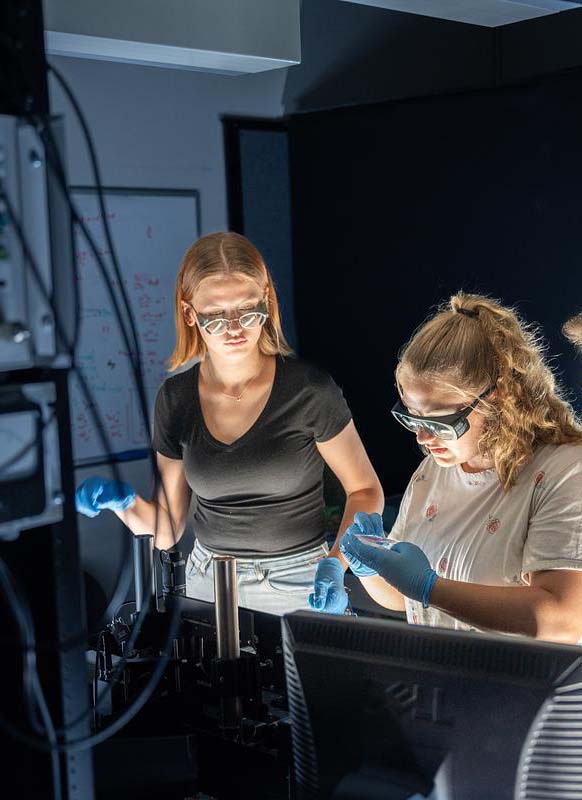 Students wearing safety glasses and gloves conducting laboratory experiment with equipment and glassware under focused lighting.