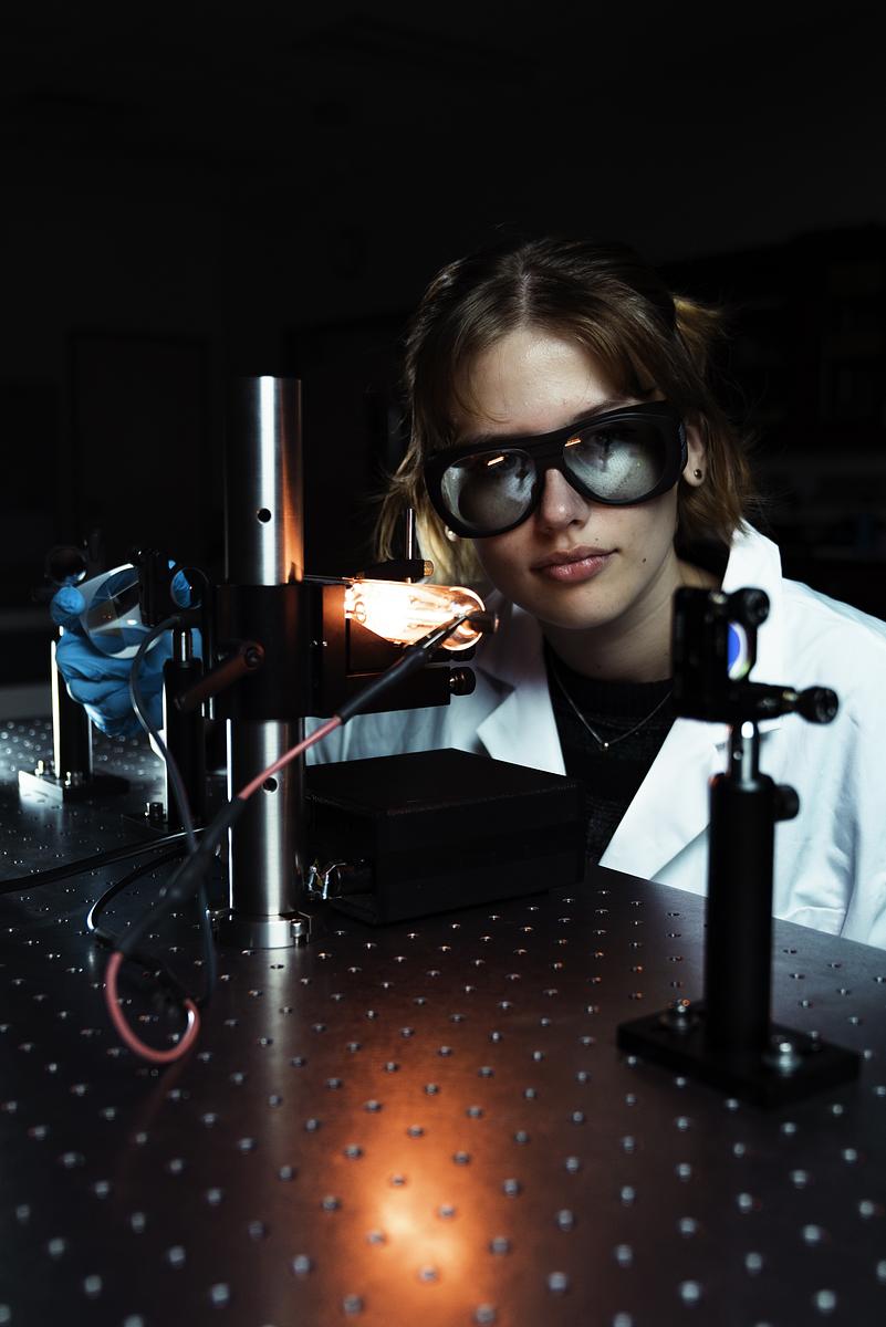 Student wearing laser safety goggles aligning optical components on photonics lab table with beam source and mounts.