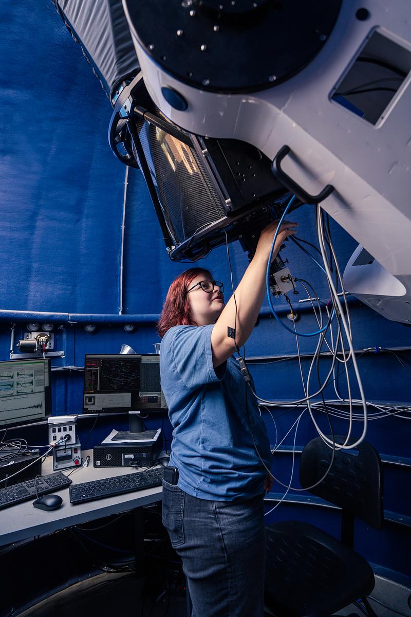 Student adjusting telescope instrument and wiring inside observatory control room with mounted optical system above.