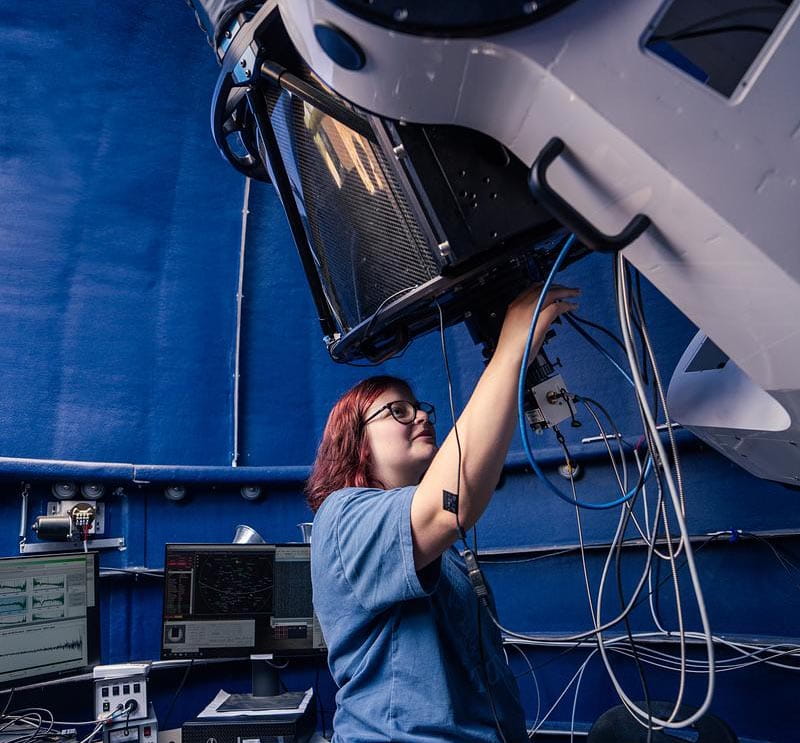 Student adjusting telescope instrument and wiring inside observatory control room with mounted optical system above.
