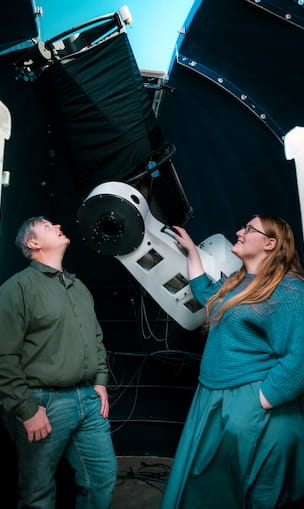 Two people standing inside observatory dome looking up out of the open shutter above.