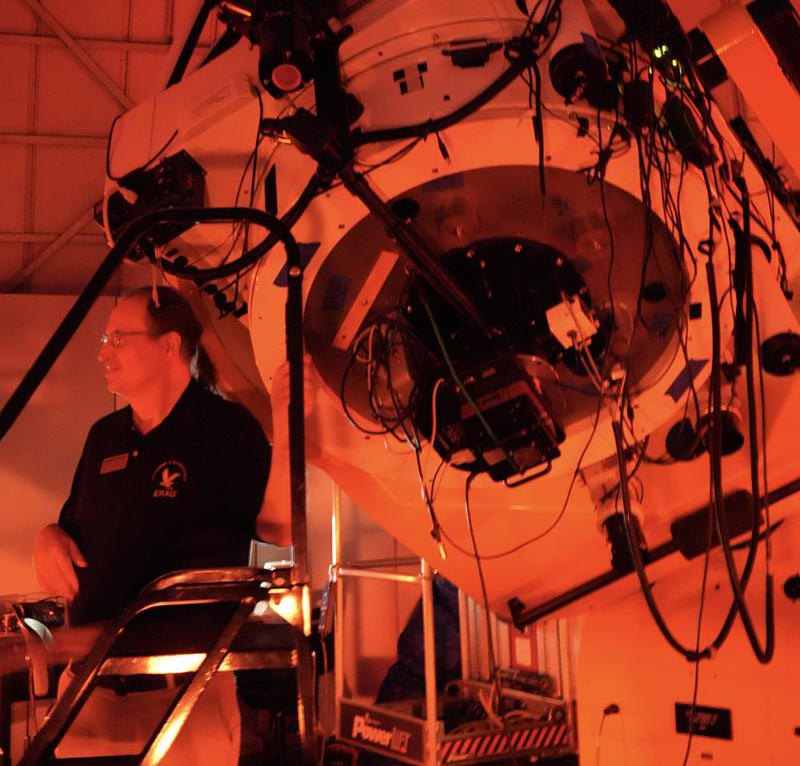 Large research telescope mounted inside an observatory dome with exposed cabling and instrumentation, as a staff member operates controls under red night-vision lighting.