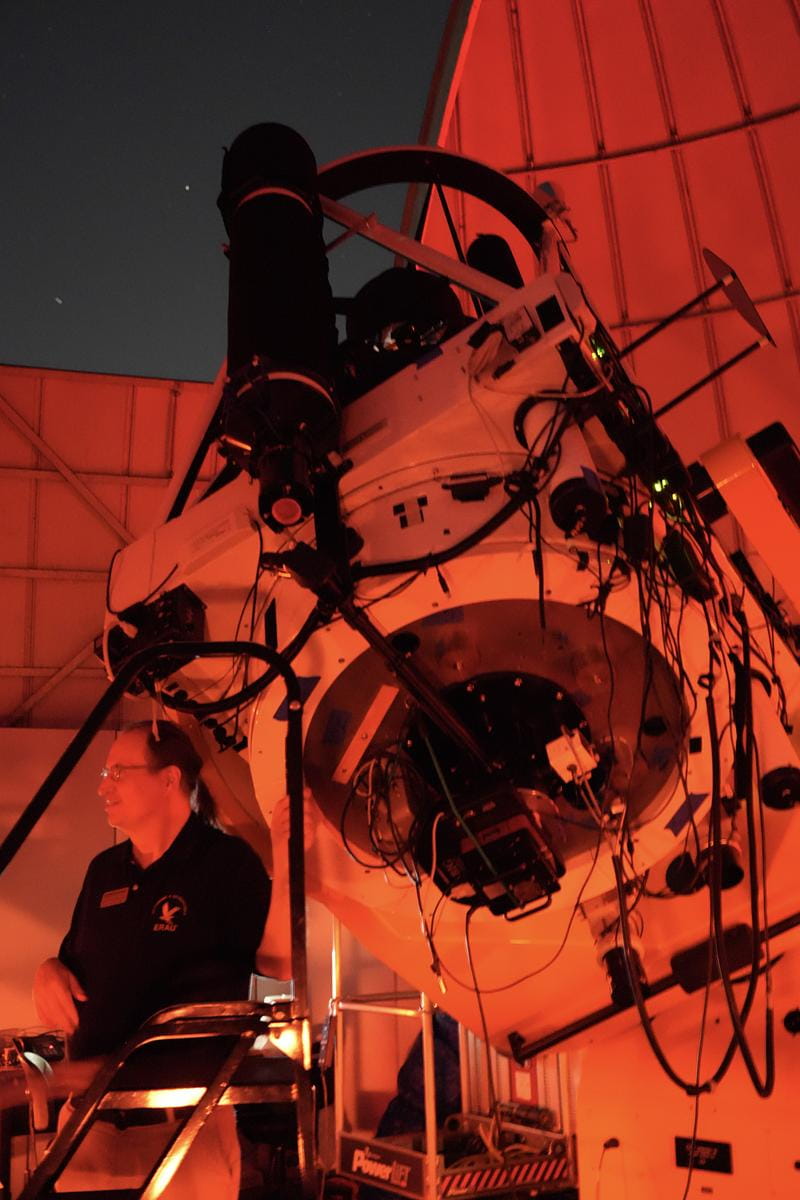 Large research telescope mounted inside an observatory dome with exposed cabling and instrumentation, as a staff member operates controls under red night-vision lighting.