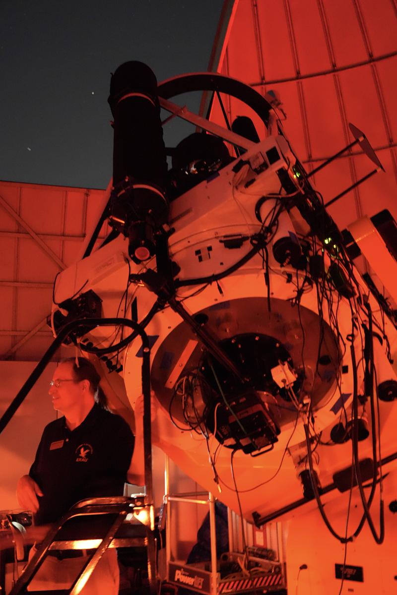 Large research telescope mounted inside an observatory dome with exposed cabling and instrumentation, as a staff member operates controls under red night-vision lighting.