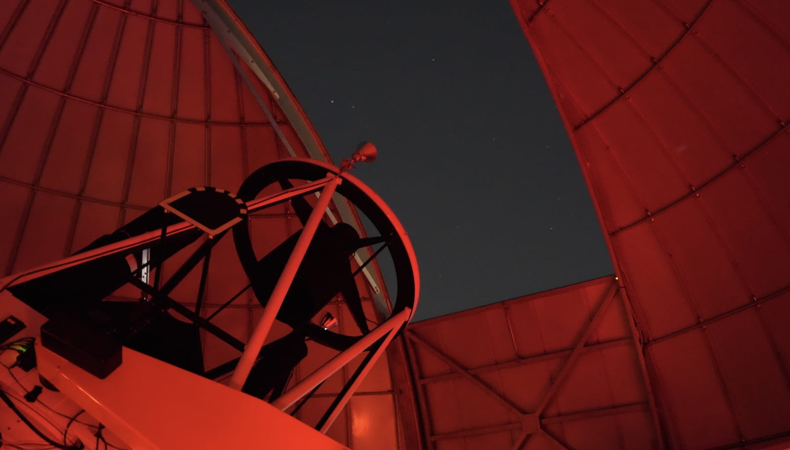 Research telescope mounted inside an observatory dome under red night-vision lighting, with the dome opening revealing a star-filled sky.