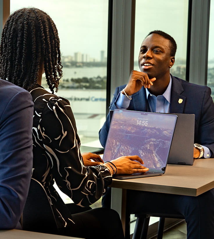 Student in business attire leads a small meeting at a table with two peers, using a laptop, seated beside large windows overlooking a coastal campus skyline and waterfront.