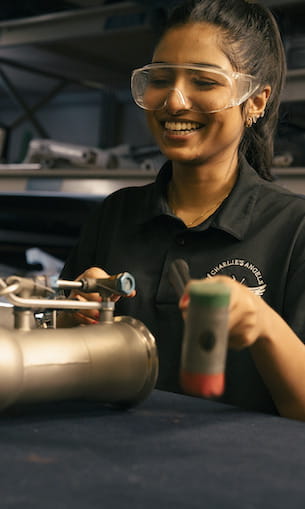 Student wearing safety goggles adjusts a cylindrical aerospace component on a lab workbench, with tools and equipment visible in the background.