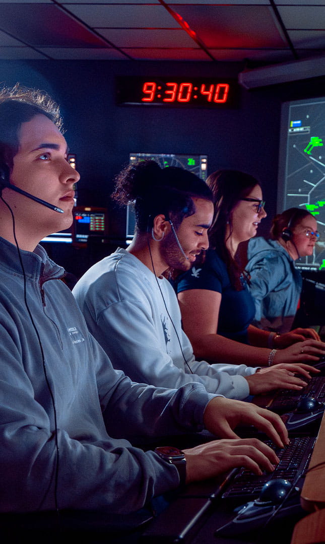 A group of people wearing headsets work at computer stations in a control room, with large screens displaying air traffic data in the background.