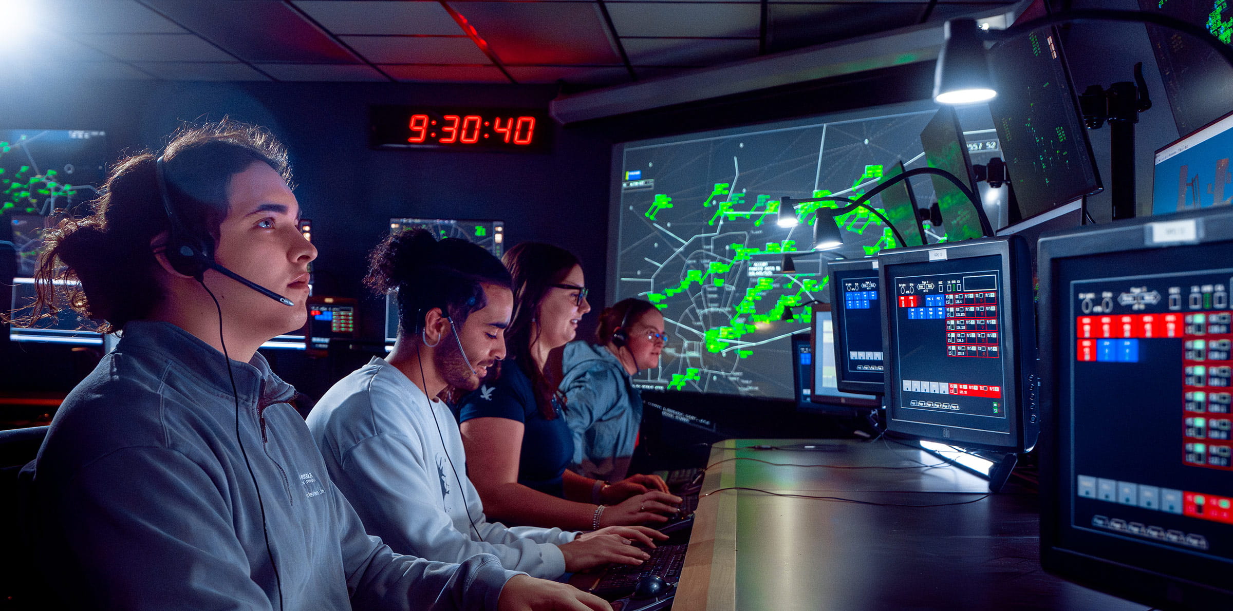 A group of people wearing headsets work at computer stations in a control room, with large screens displaying air traffic data in the background.