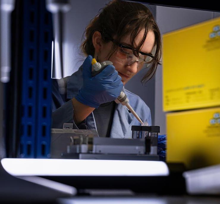 Student wearing safety glasses and nitrile gloves uses a micropipette to transfer liquid into a laboratory sample container at a research bench.