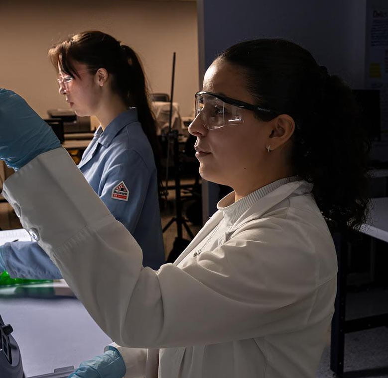 Students wearing lab coats, safety glasses, and gloves prepare samples with pipettes and a benchtop centrifuge in a biomedical laboratory.