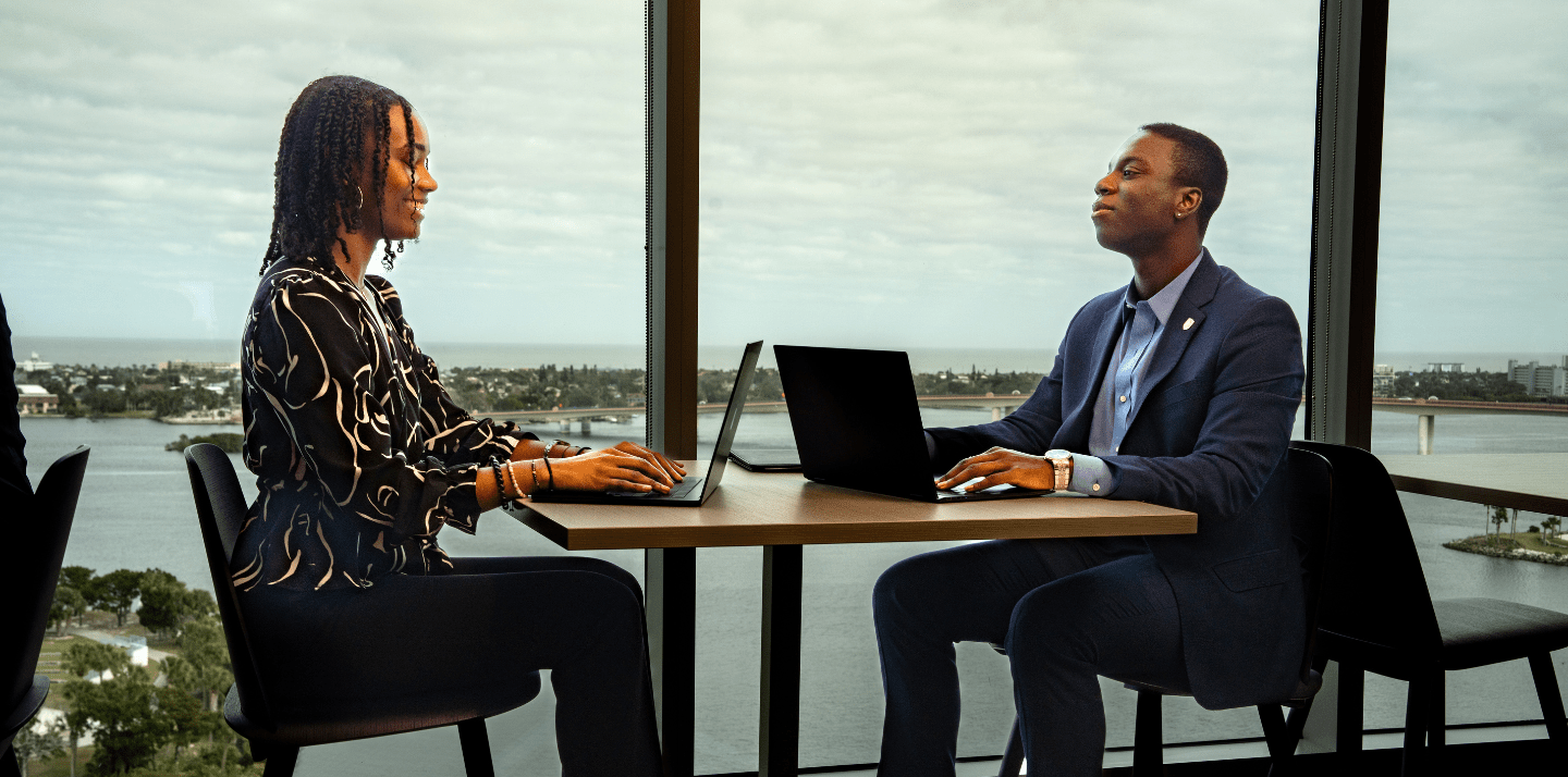 Two students seated across a small table using laptops, facing each other in a high-rise campus study space with floor-to-ceiling windows overlooking a coastal waterway and bridge.
