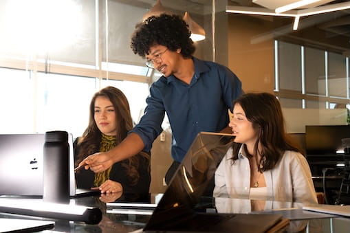 Students collaborating at computer workstation, one pointing at screen while others review work in modern office setting.
