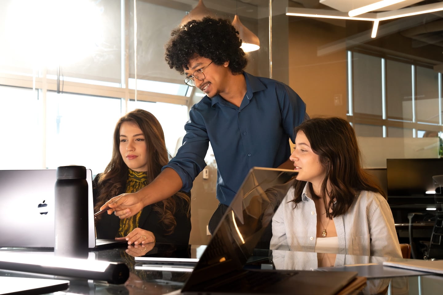 Instructor points at a laptop screen while two students sit at a conference table with open laptops, glass-walled classroom with pendant lighting and large windows in the background.