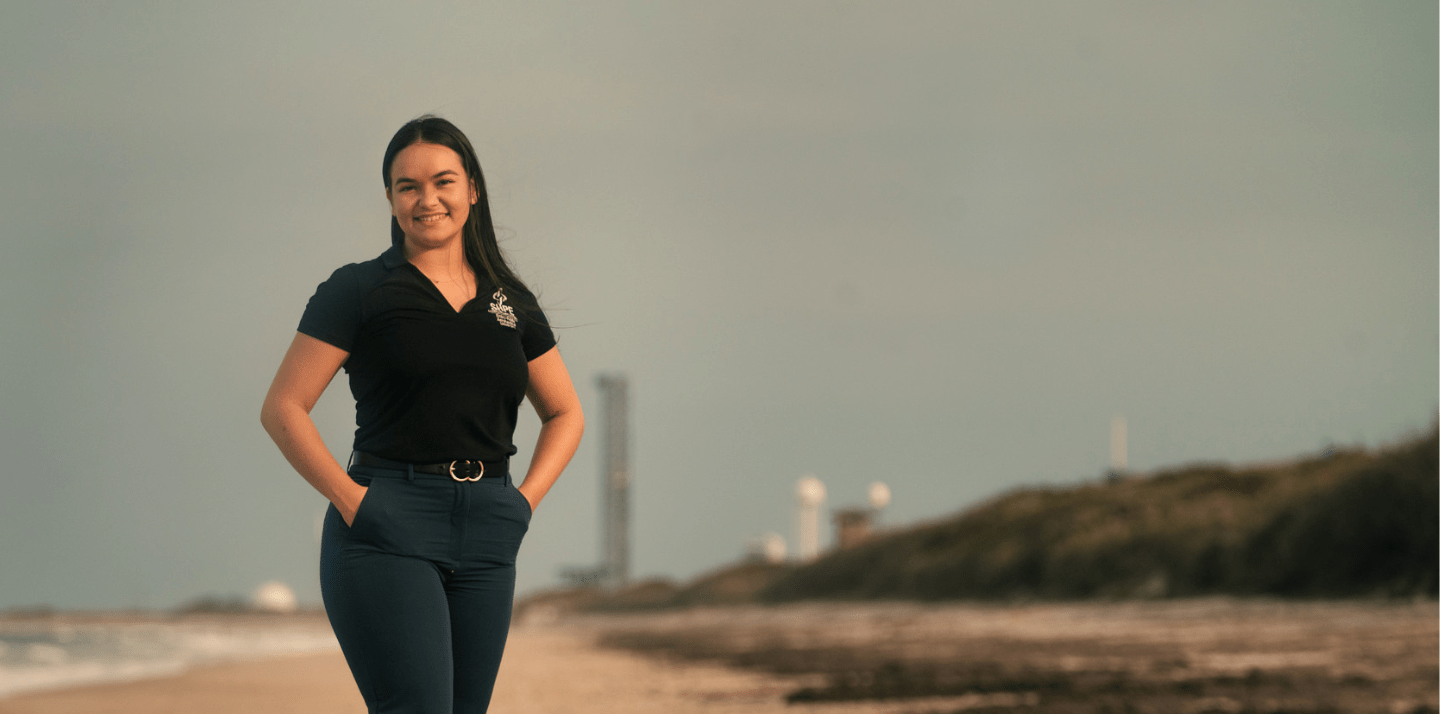 Student stands on a beach near a coastal launch complex with launch tower and radar domes visible in the background.