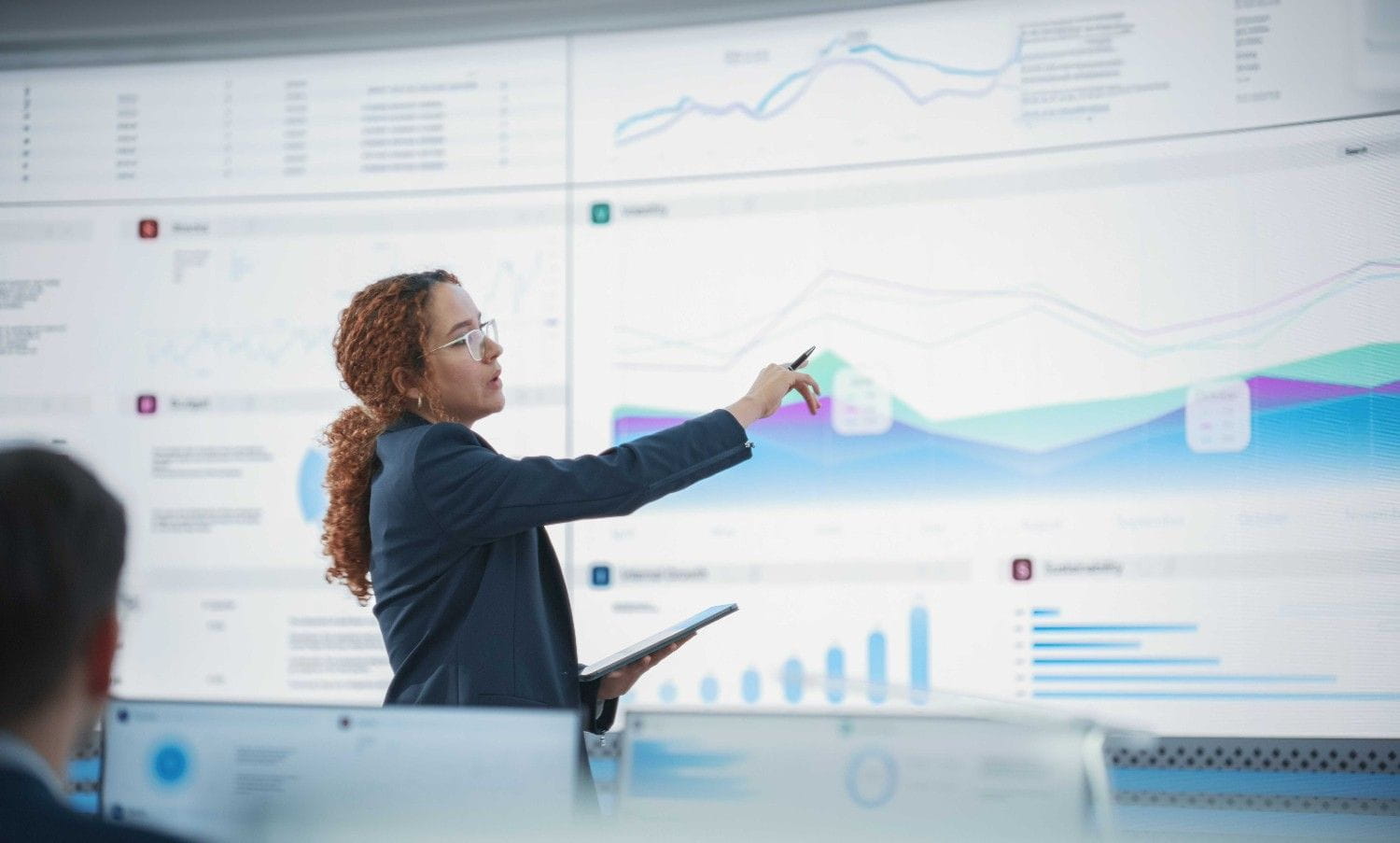 Instructor presents data visualizations and trend graphs on a large interactive display while holding a tablet in a business analytics classroom.