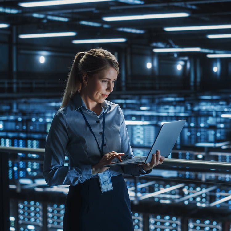 Woman using a laptop in data center environment with rows of servers and network equipment.