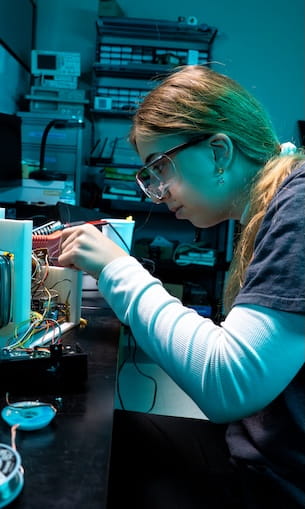 Student wearing safety glasses assembling electronic components and wiring on open device in lab.