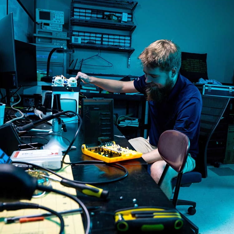 Student assembles and tests a small robotic device with human-looking eyeballs at a lab workbench with tools, soldering equipment, and circuit components.