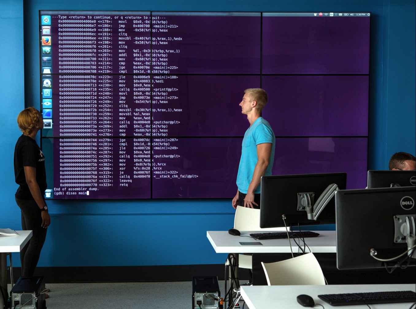 Students review assembly code displayed on a large multi-screen video wall in a computer science classroom with desktop workstations in the foreground.