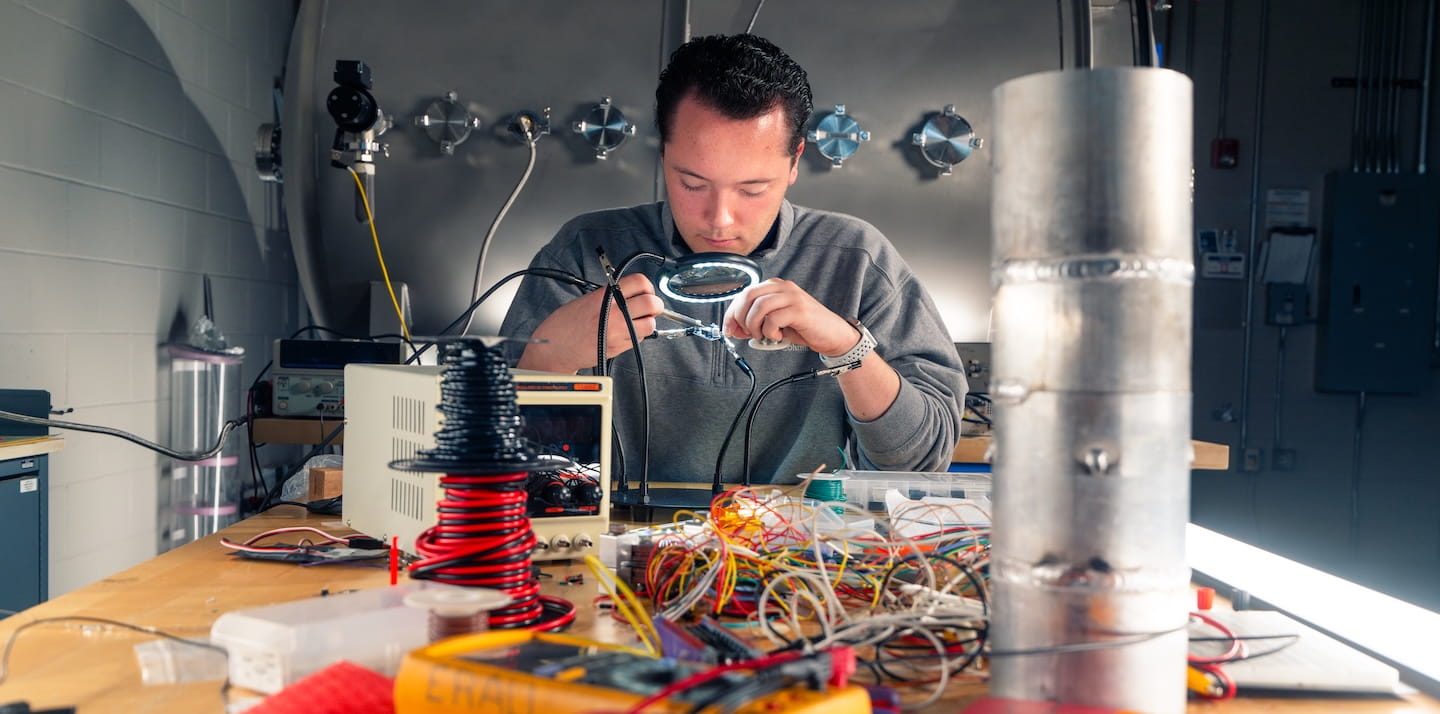 Student uses a magnifying lamp to solder electronic components on a circuit board at a workbench with oscilloscopes, wiring, and a high-voltage coil in an engineering lab.