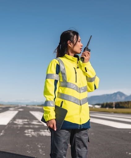 A person in a yelow high-visibility jacket stands on an airport runway holding a walkie-talkie. 