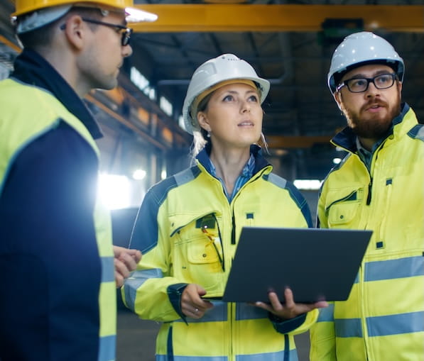 Three people in high-visibility jackets and hard hats on a worksite. One person is holding a laptop computer.