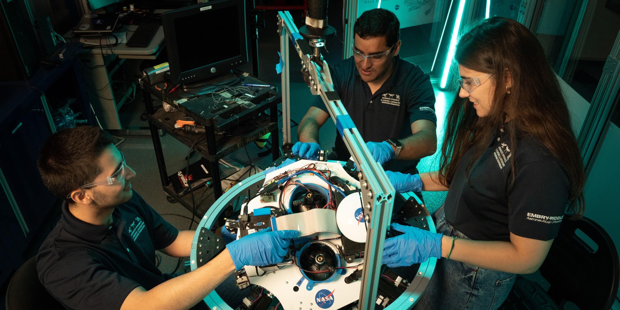 Three students in safety glasses and gloves assemble a circular NASA robotic system with exposed wiring, motors, and sensors on a lab workbench, using an aluminum frame test rig and nearby computer equipment in an engineering lab.