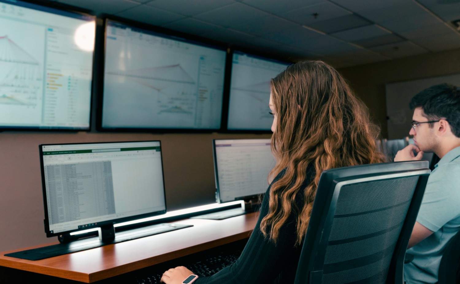 Students analyze data on desktop monitors displaying spreadsheets and charts, with multiple large screens showing trend graphs in a data analytics lab.