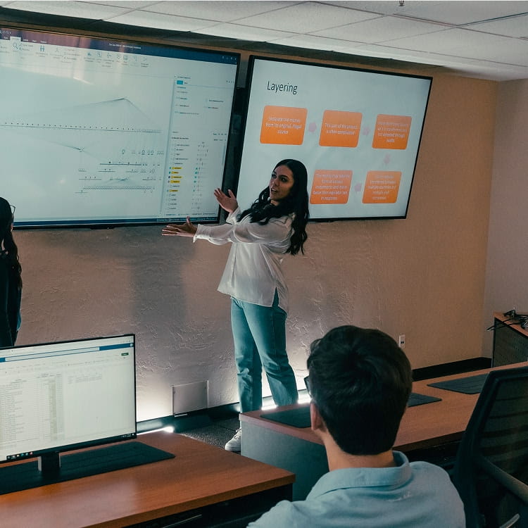 Student presenter gestures toward two wall-mounted displays showing a data visualization timeline and a slide titled Layering with process steps, classmates seated at desktop workstations in a classroom.