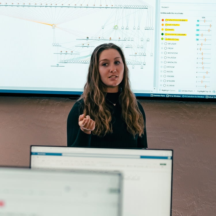 Student presenter gestures while explaining a network data visualization with nodes and links displayed on a large wall-mounted screen, classmates’ monitors visible in the foreground.