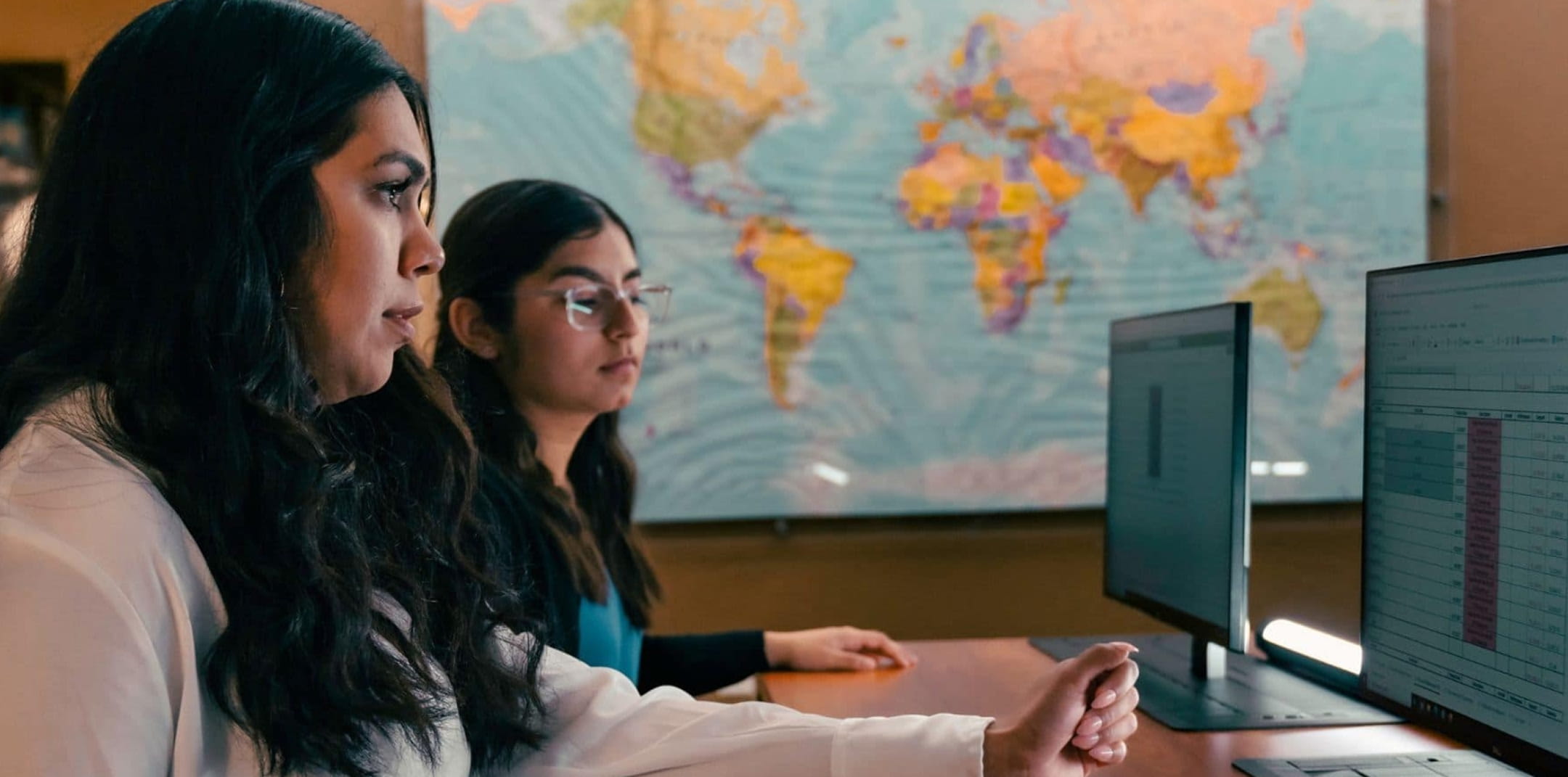 Two students review data on dual desktop monitors displaying a spreadsheet, seated at a desk with a large world map mounted behind them.