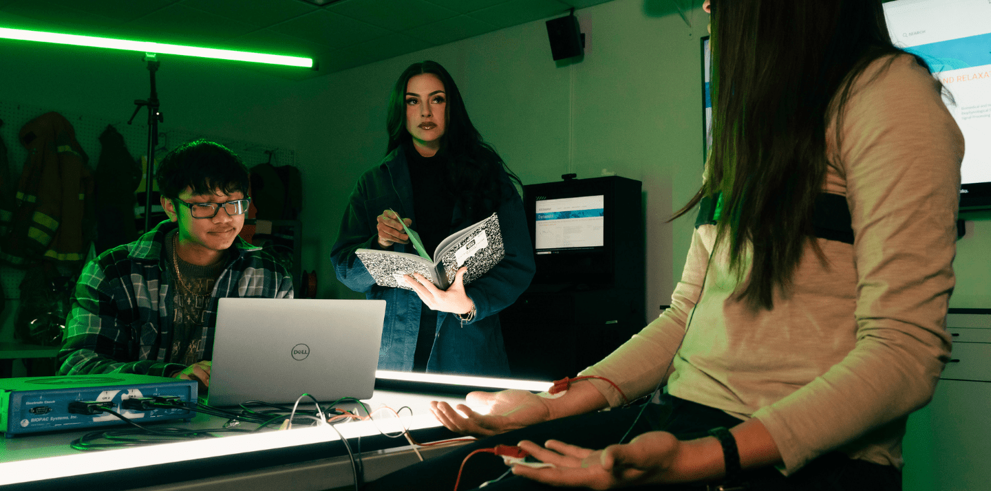 Students conduct a bioinstrumentation experiment with electrodes attached to a participant’s arm while monitoring data on a laptop and laboratory equipment under green accent lighting.
