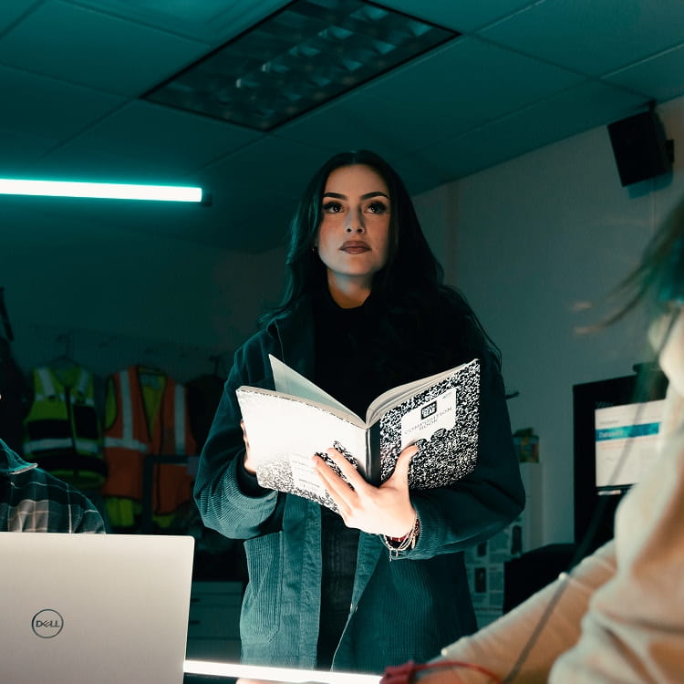 Student stands reading from an open composition notebook while addressing classmates at desks with laptops, safety vests hanging on the wall behind in a lab-style classroom.