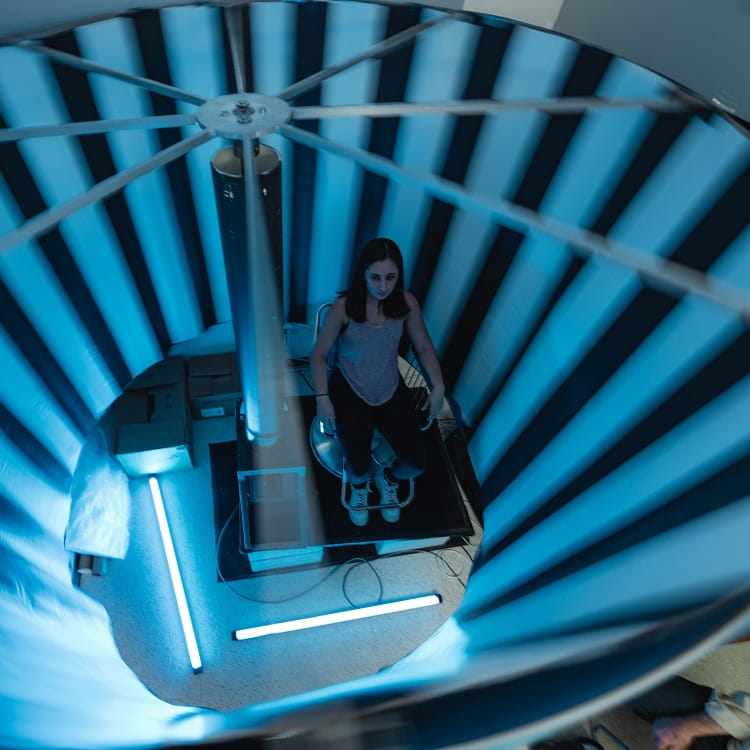 Student stands on a platform inside a cylindrical test chamber with radial panels and a central column, likely an acoustic or electromagnetic testing setup, viewed from above.