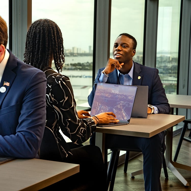 Student in business attire leads a small meeting at a table, using a laptop, seated beside large windows overlooking a coastal campus skyline and waterfront.