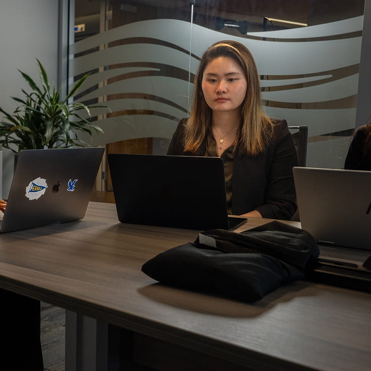 Student works on a laptop at a conference table with two additional laptops nearby, glass wall with frosted wave pattern behind, office meeting room setting.