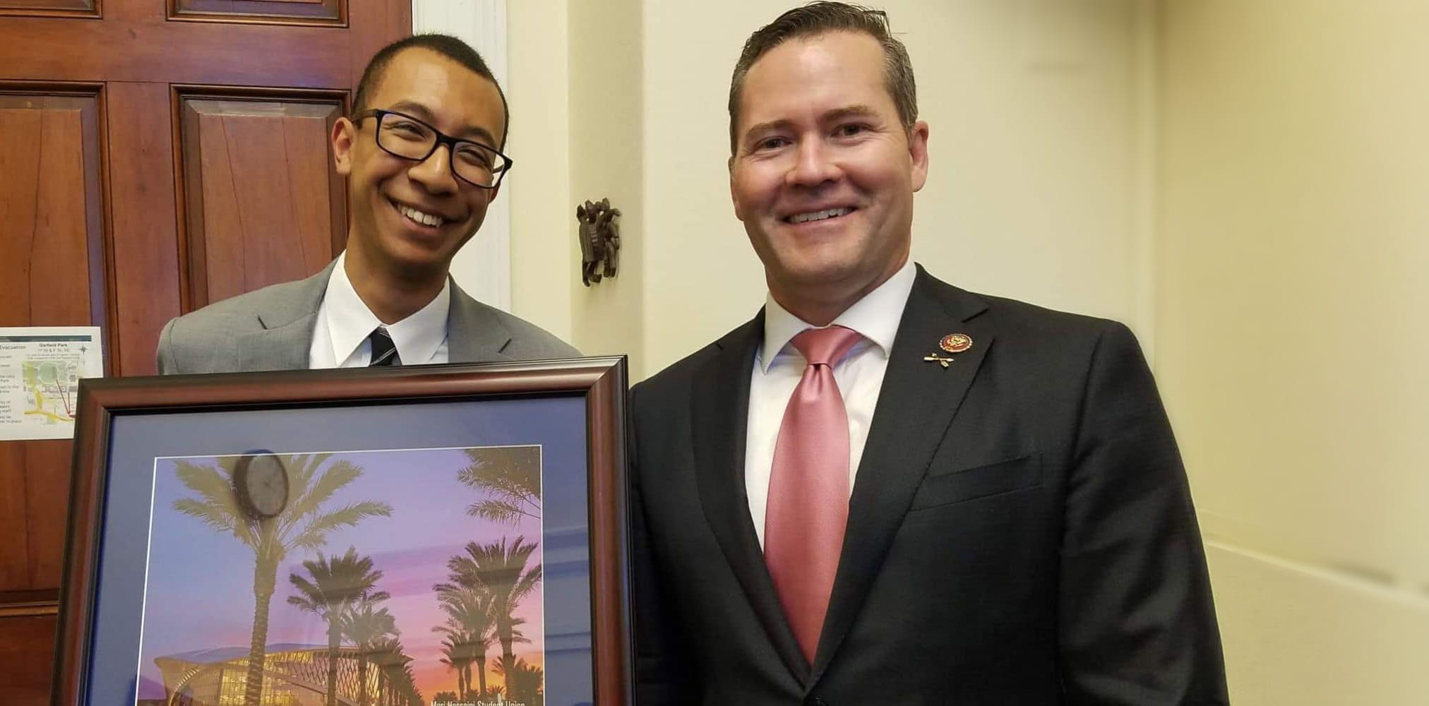 Two men in suits hold a framed image of a palm-lined campus entrance with a modern building at sunset, posed indoors near a wooden door.