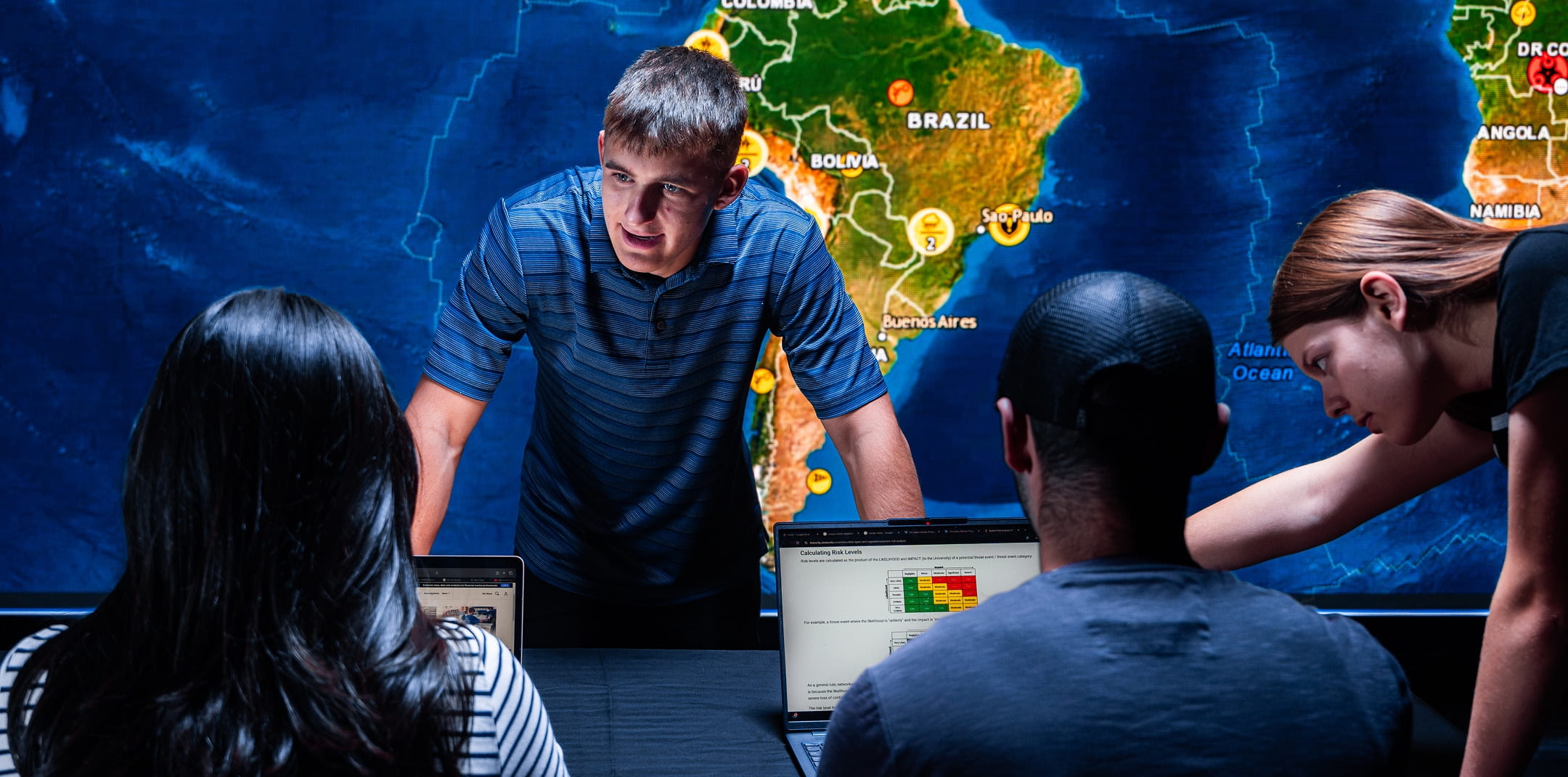 Students collaborating at a table with laptops displaying a risk analysis chart, large digital world map focused on South America with location markers behind them, one student leaning forward leading discussion.