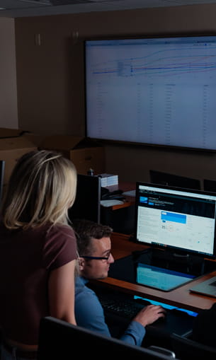 Two individuals collaborate at a computer in a classroom-style environment.