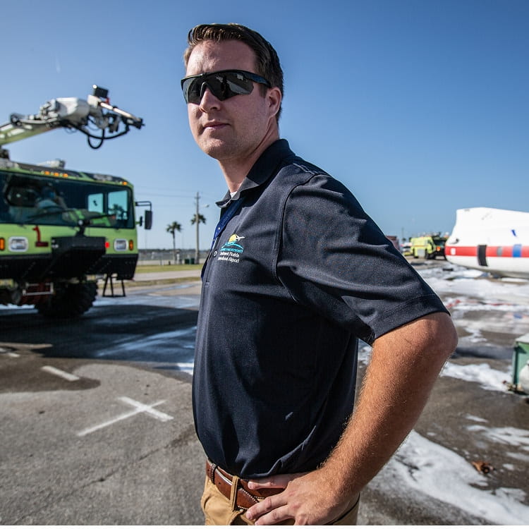 Student in safety sunglasses and Embry-Riddle polo stands on airport training ramp near ARFF crash truck spraying foam, with aircraft fuselage trainer and emergency vehicles in background.