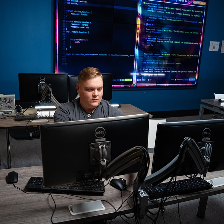 Student works at a multi-monitor computer station with large wall displays showing source code and software development tools in a computer science lab.