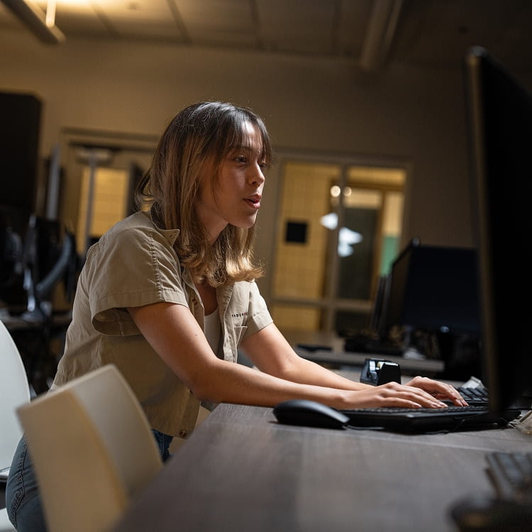Student working at a desktop computer workstation in a campus computer lab, typing on a keyboard with focused posture; rows of monitors and lab equipment in the background.