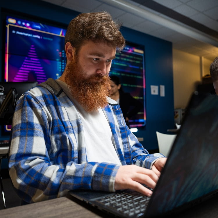 Student coding on a laptop in a computer lab, large wall displays behind show colorful code and data visualizations; other students and workstations in the background.