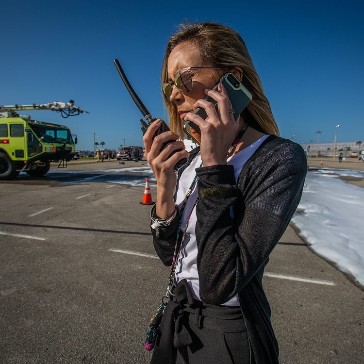 Airport operations staff member coordinating via handheld radio and smartphone on an aircraft ramp, fire rescue truck and foam on pavement behind, traffic cone and runway markings visible.