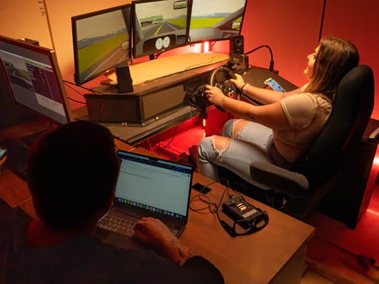 One person operates a driving simulator as another observes.