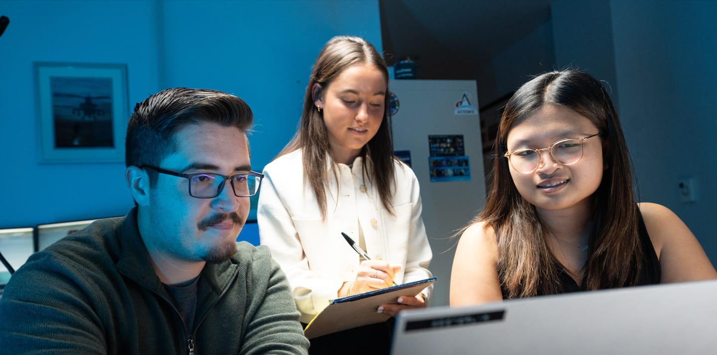 Three people collaborate around a laptop while one person takes notes on a notepad.