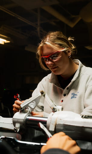 A person wearing safety goggles works with tools on a mechanical assembly in a workshop.
